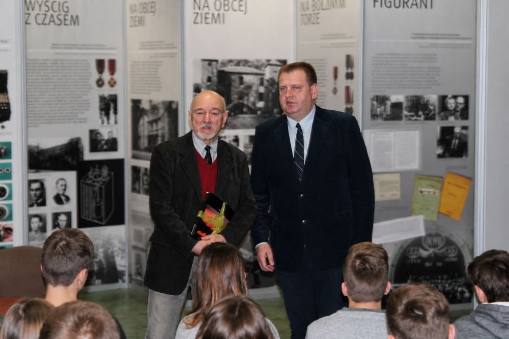 Dwóch mężczyzn przed grupą młodzieży na tle wystawy posterowej.