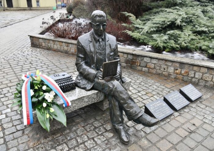 Pomnik Mariana Rejewskiego w formie ławeczki, na której sieci postać. Obok wieniec z szarfami.