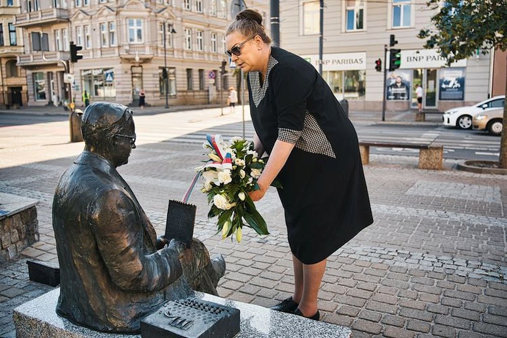 Kobieta z wiązanką kwiatów pochyla się nad pomnikiem-ławeczką.